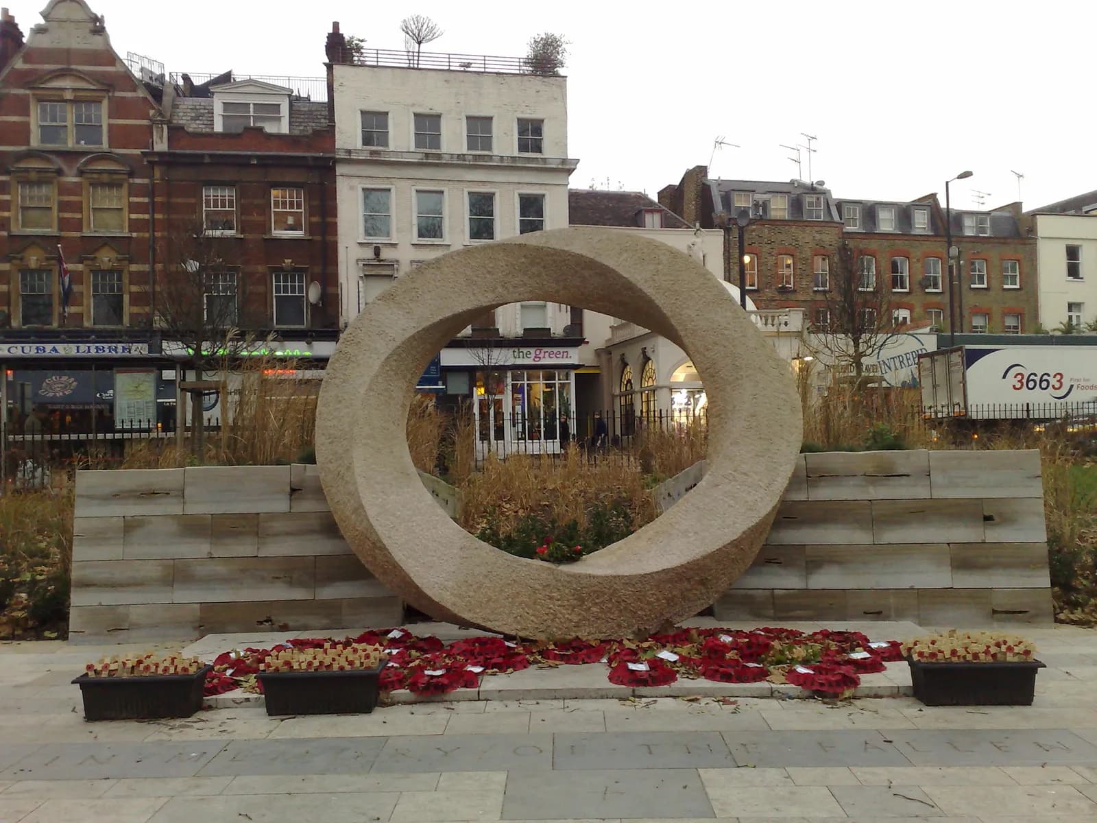 A large stone Mobius band sculpture stands prominently in the center of an urban square, surrounded by numerous red wreaths and potted plants, with historic buildings and storefronts in the background under an overcast sky.