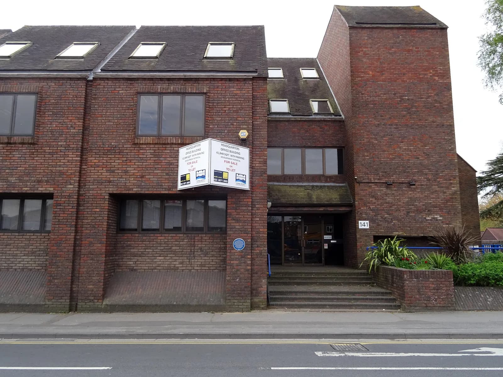 A red brick office building with several windows and roof lights features a 'for sale' sign hanging on the front.