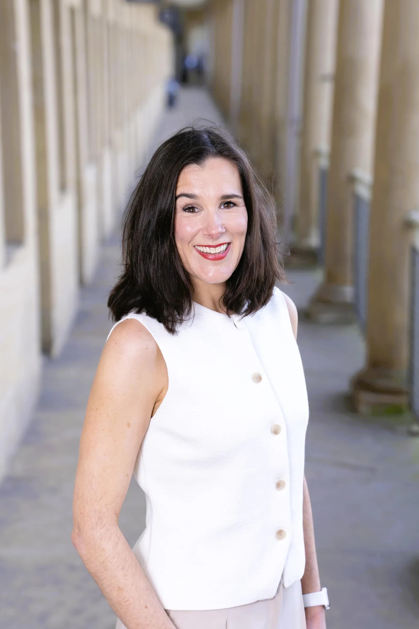 A smiling woman with dark hair and a white vest stands in a long, sunlit hallway lined with pillars.