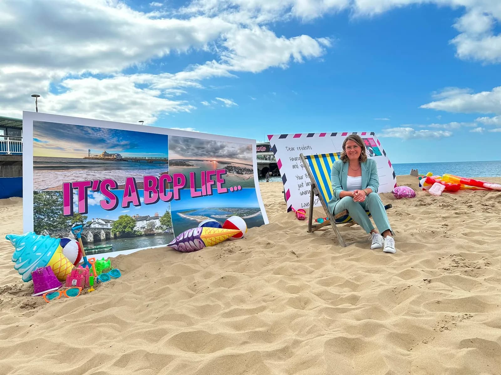 A woman smiles while sitting in a beach chair at an outdoor event on a sandy beach, with a large billboard display and postcard cutout behind her.