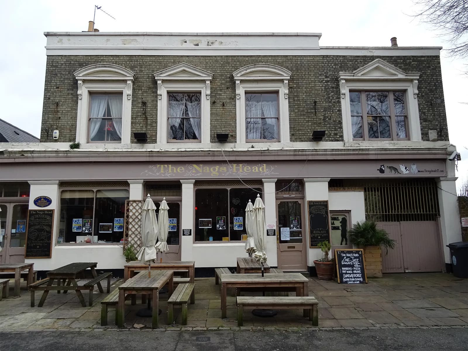 A two-story building with a brick facade houses a pub called "The Nags Head," featuring outdoor picnic tables and umbrellas on a paved patio.