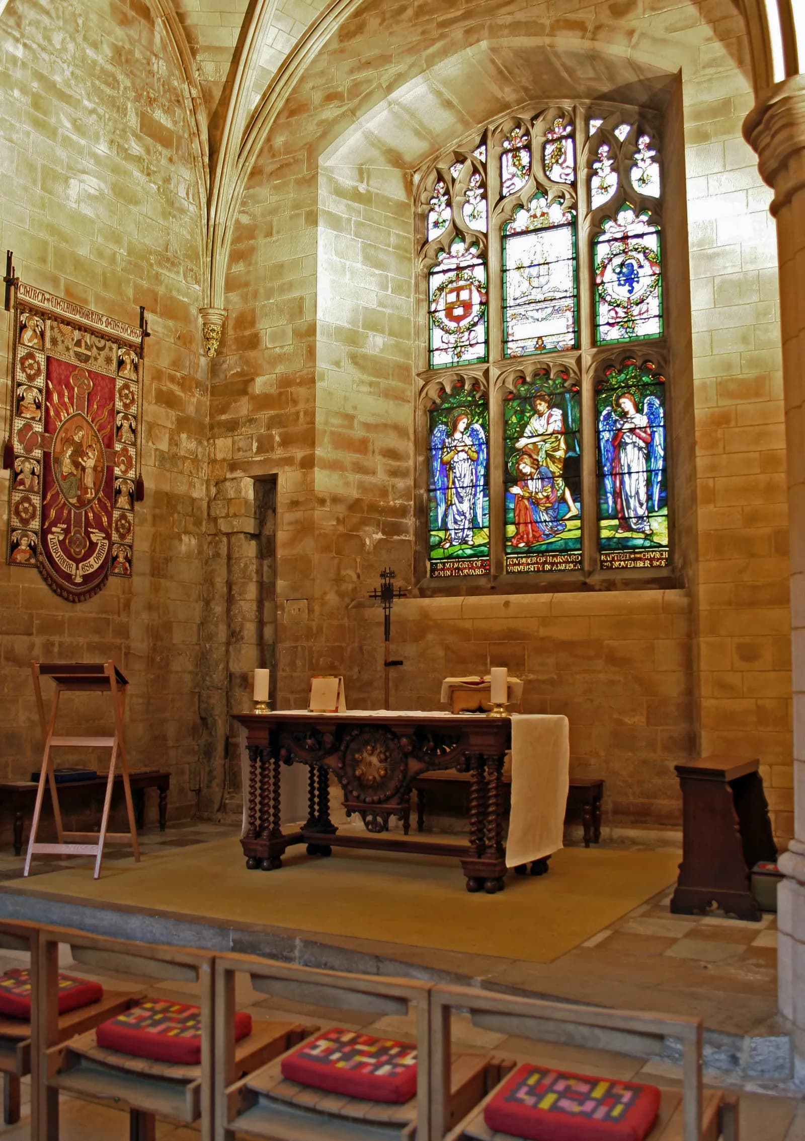 A historic church interior features a carved wooden altar table, an ornate tapestry, and a large stained-glass window depicting religious figures in vibrant colors.