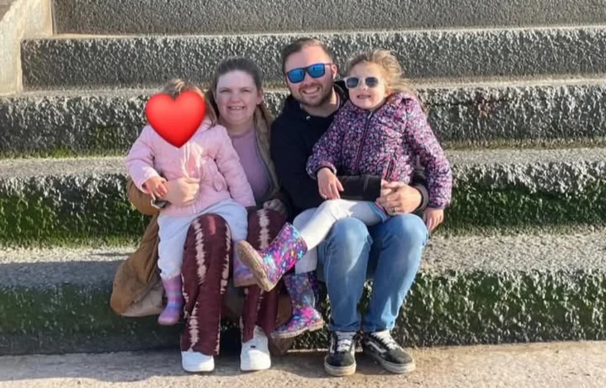 A family of four, including two young girls, a woman, and a man, sit together on stone steps, all smiling at the camera.