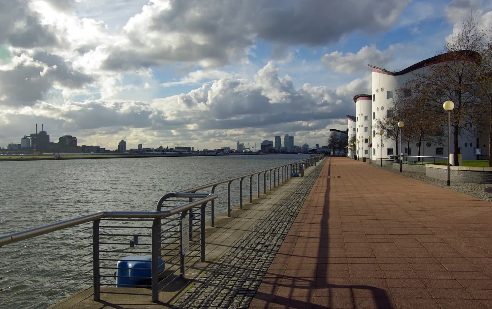 A scenic view of a waterfront promenade with modern white buildings and trees on the right, calm water in the middle, and a distant city skyline under a partly cloudy sky.