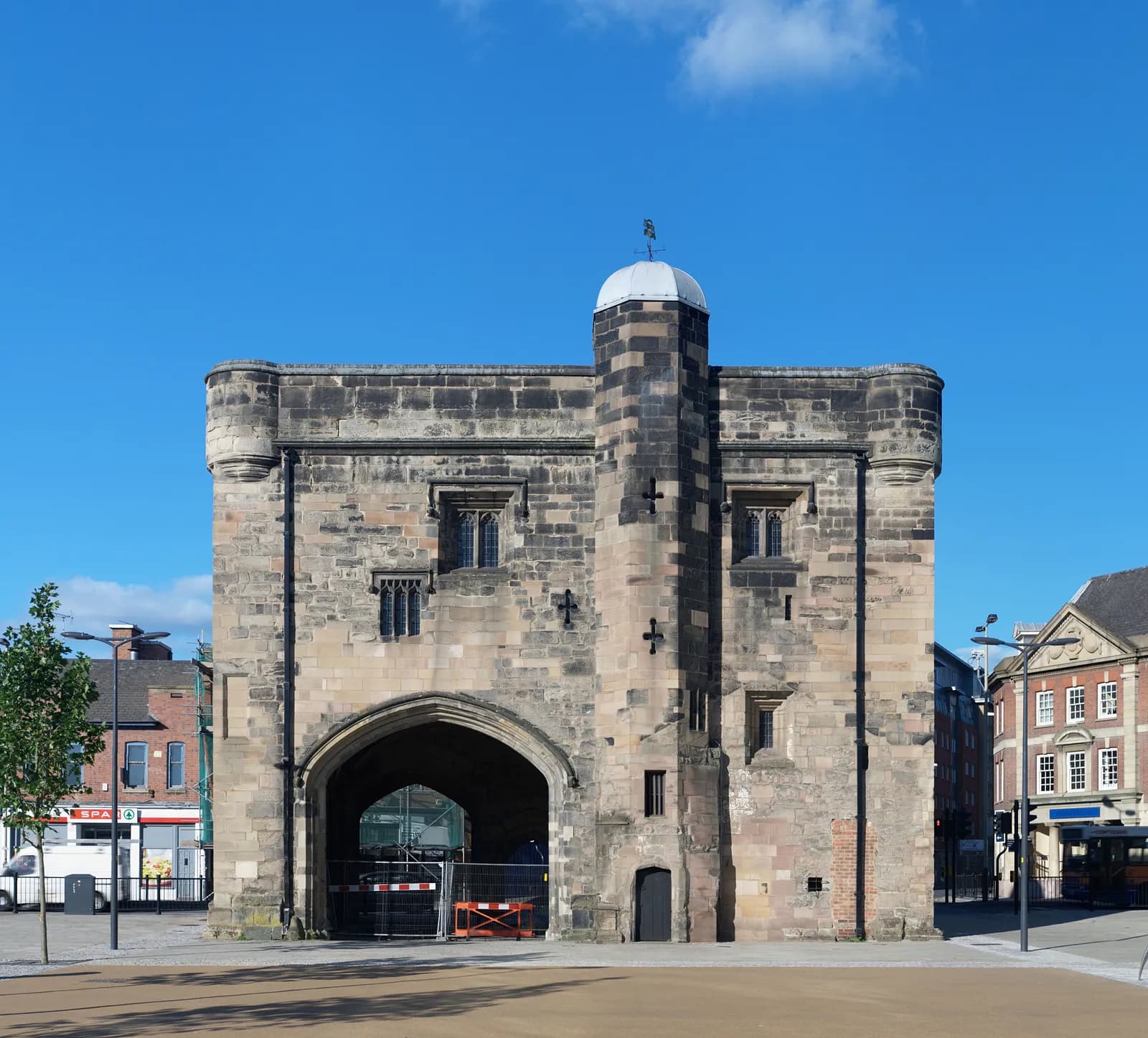 A historic stone gatehouse with an arched passageway and a small tower, seen on a sunny day with clear blue sky, with modern buildings visible around it.