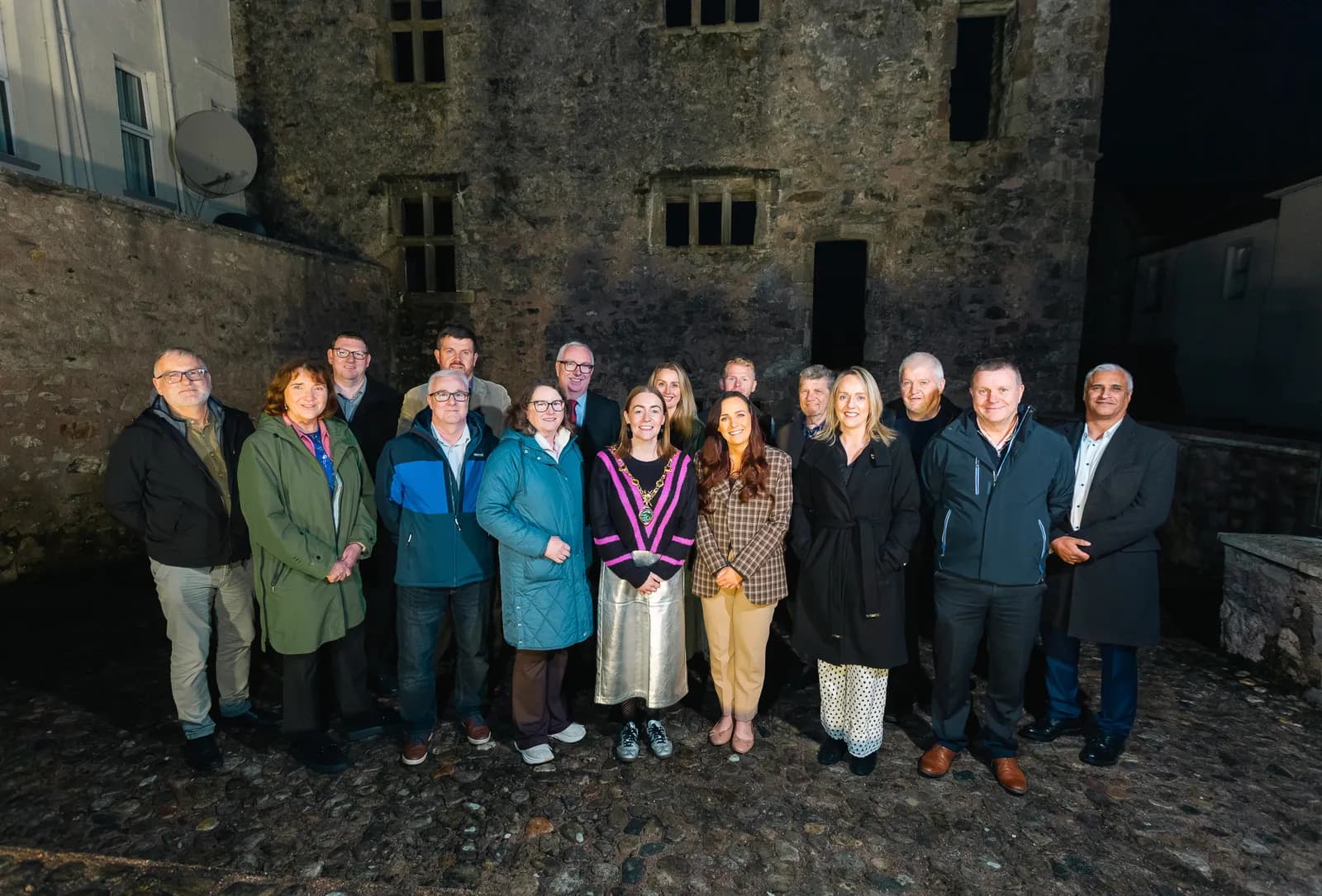 A group of eighteen people, including one woman wearing a ceremonial chain, pose for a photo in front of a stone building at night.