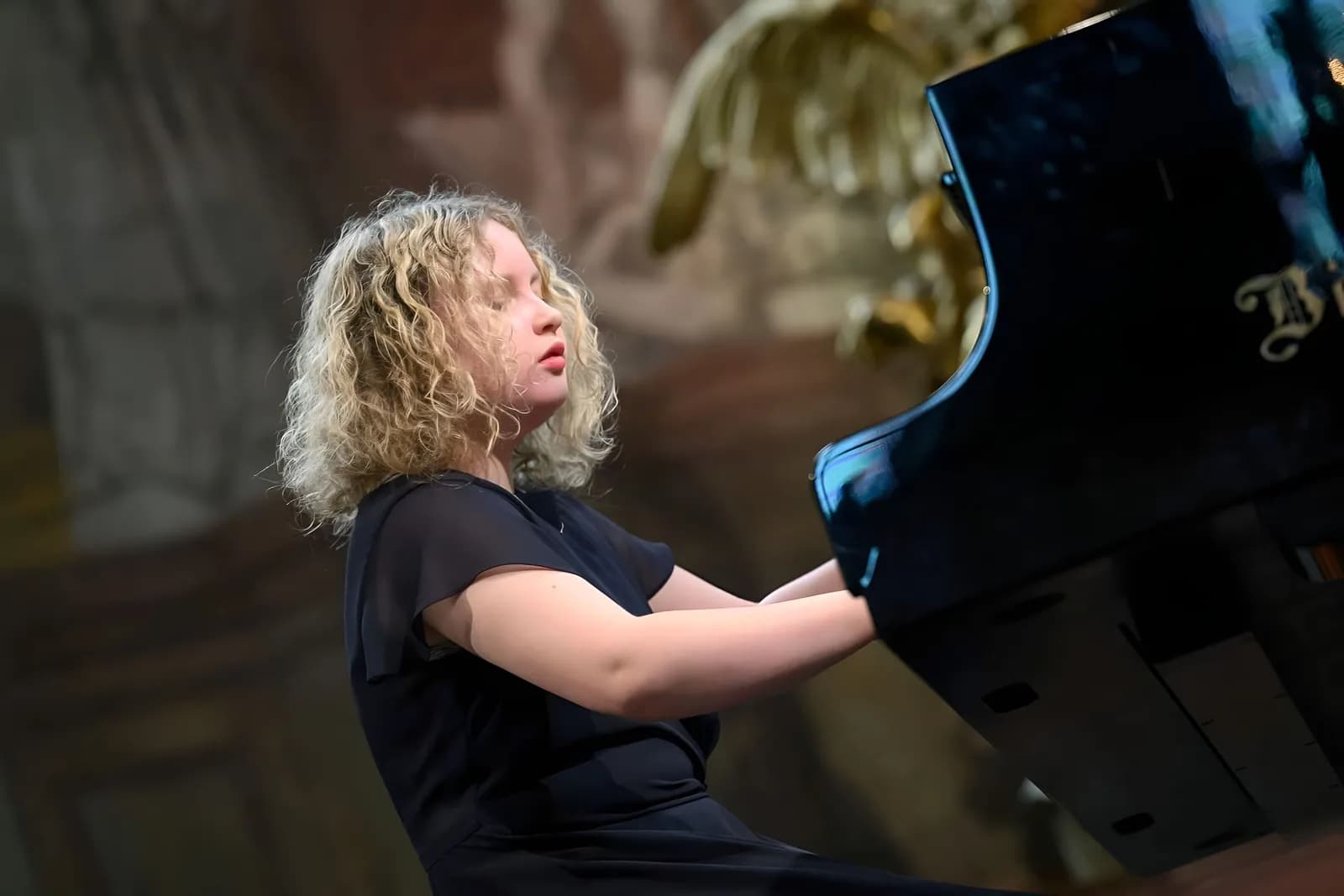 A woman with curly blonde hair in a black dress playing a grand piano with eyes closed, lost in music.