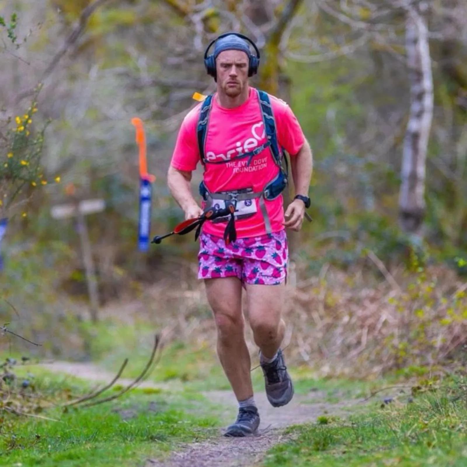 A man wearing a pink shirt, strawberry-patterned shorts, and headphones runs on a dirt path through a wooded area.