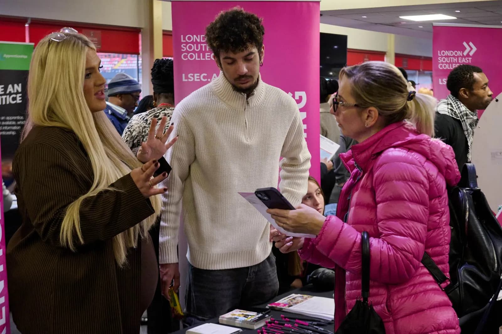 Three people engage in conversation at what appears to be a college fair or event, with two women and one man standing amongst promotional banners.