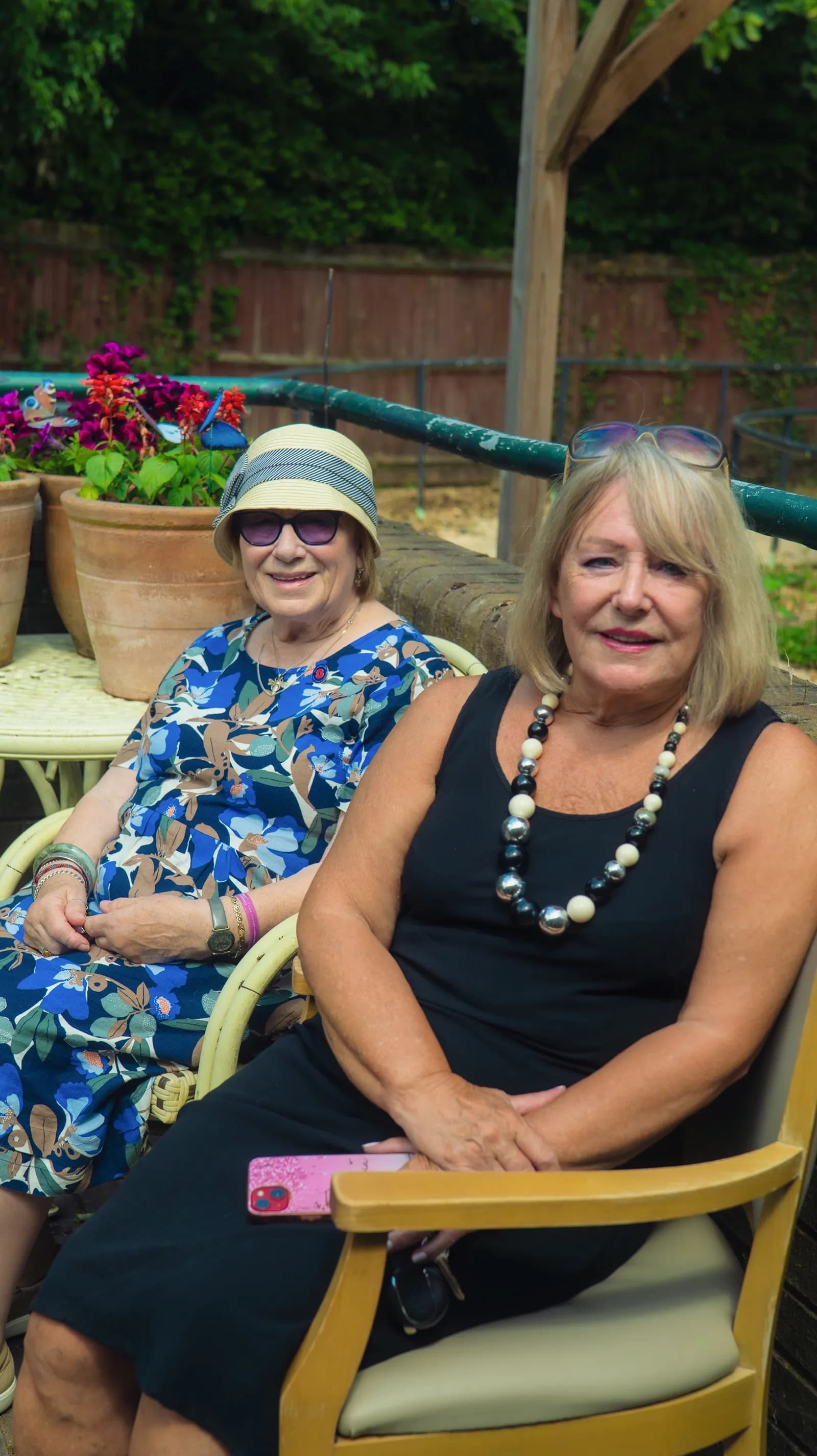 Two smiling women seated outdoors, one in a blue patterned dress and sun hat, the other in a black dress with a beaded necklace.