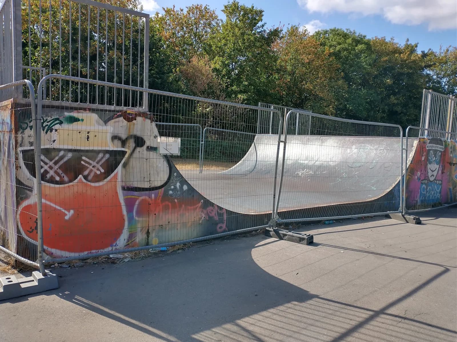 A fenced-off skate ramp covered in colorful graffiti, with trees and a blue sky in the background.