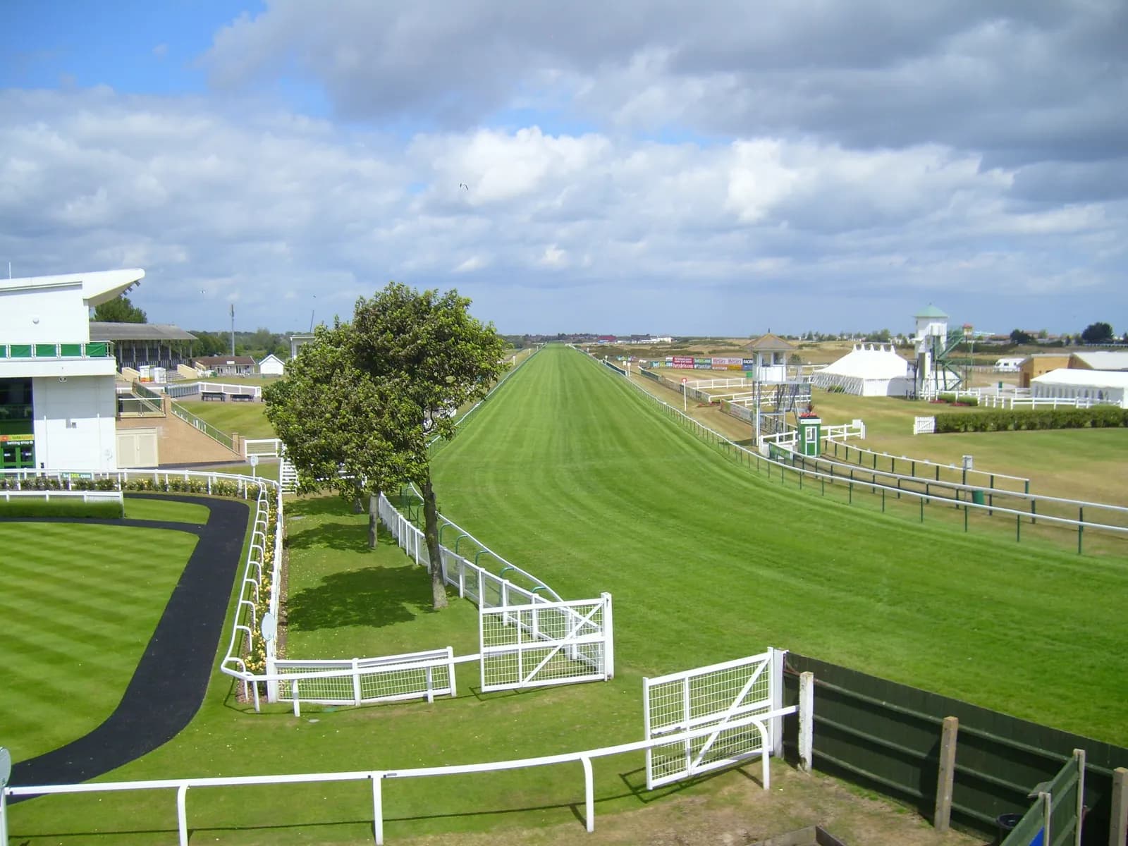A wide view of an empty green horse racing track under a partly cloudy sky, with grandstands and tents visible in the background.