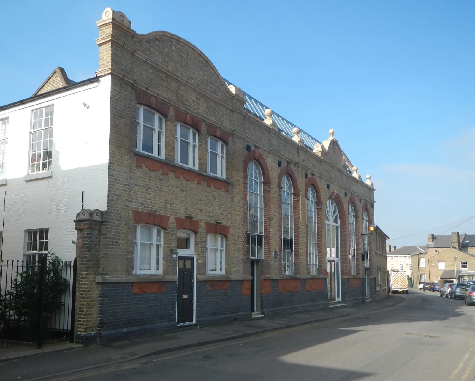 A long, ornate brick building with tall arched windows and a white building attached on the left, situates along a quiet street with parked cars.