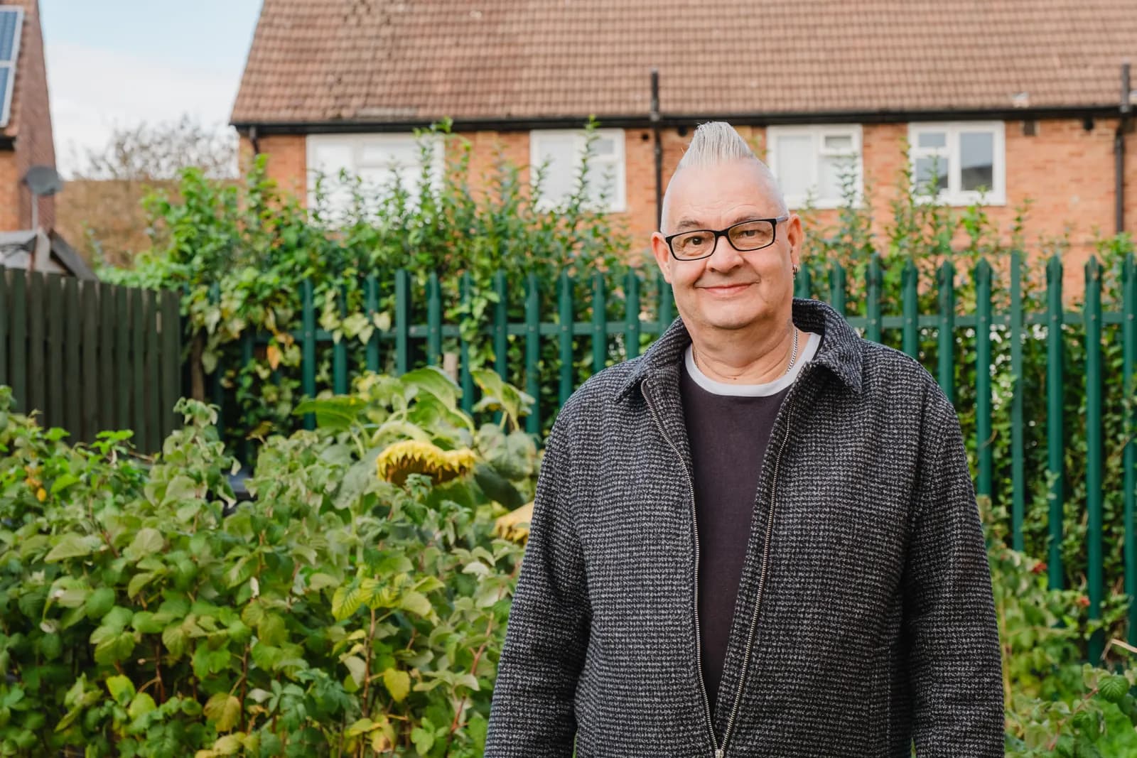 A smiling man with short white spiky hair and black glasses stands in a garden with green plants and a fence behind him, with brick houses in the background.