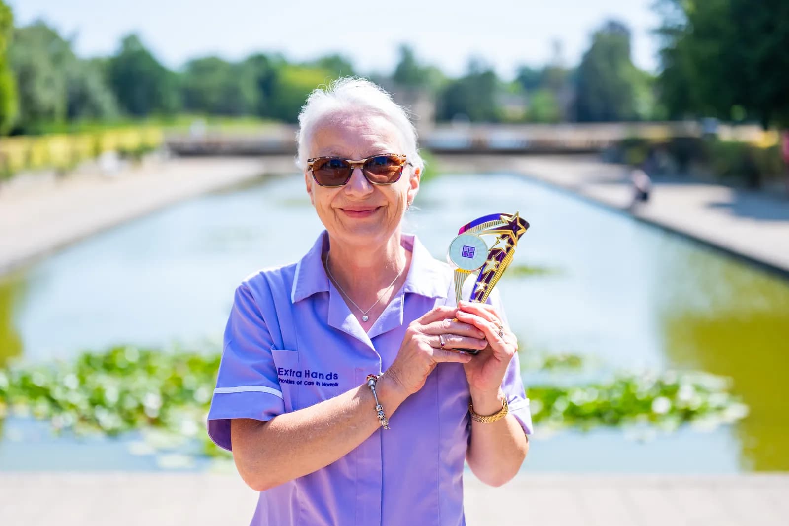 A smiling woman with white hair and sunglasses, wearing a purple uniform with 'Extra Hands' embroidered on it, holds a trophy while standing outdoors by a water feature.