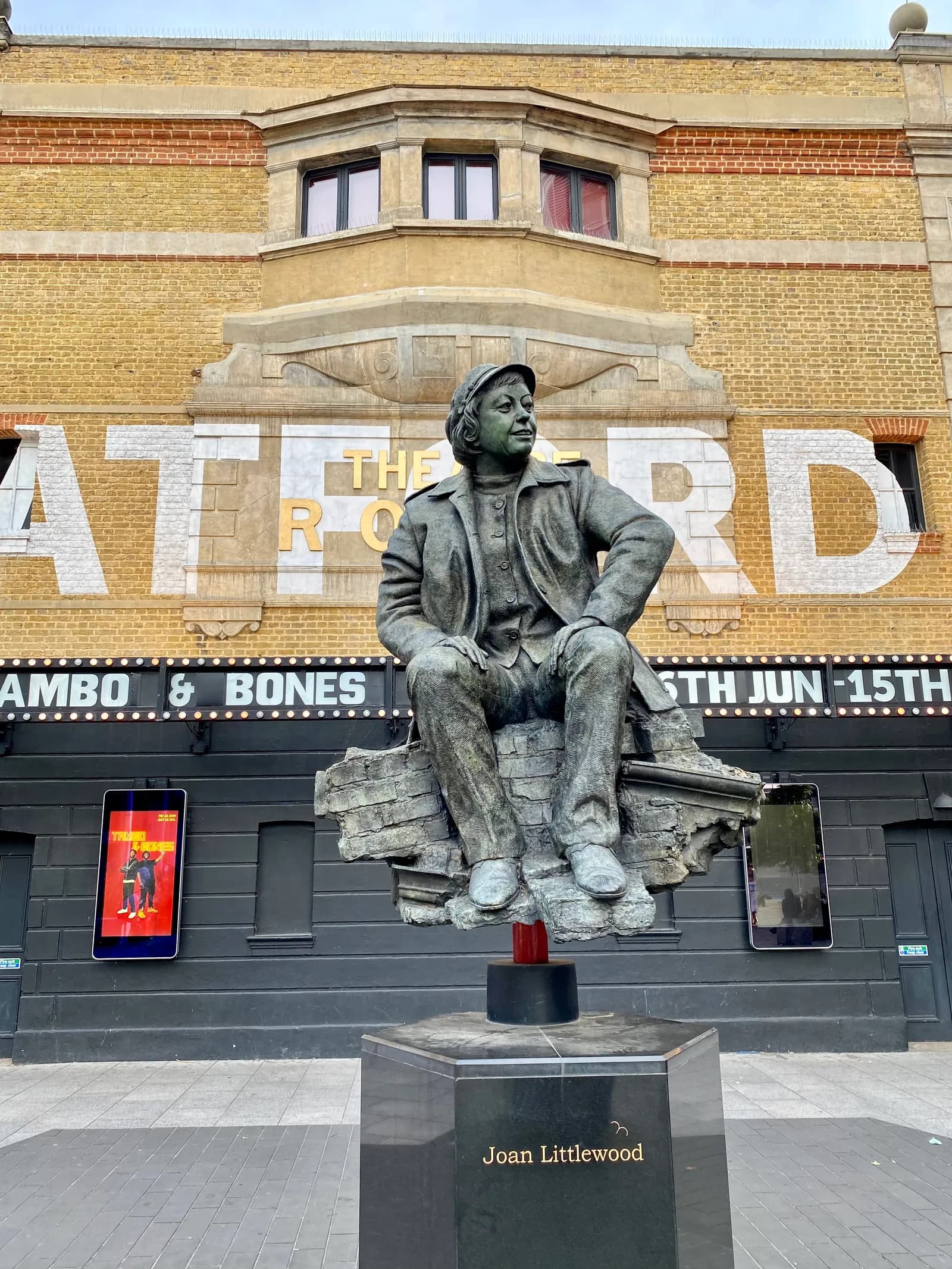 A bronze statue of Joan Littlewood sits in front of a brick building with white lettering, spelling out "THE RO".