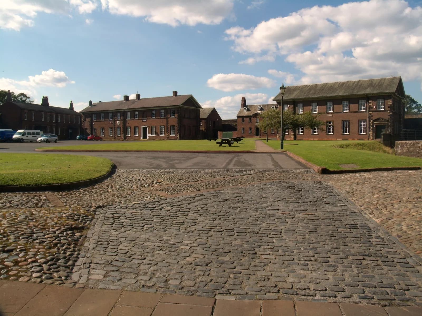A courtyard with cobblestone paths, green lawns, and several large brick buildings under a partly cloudy sky.