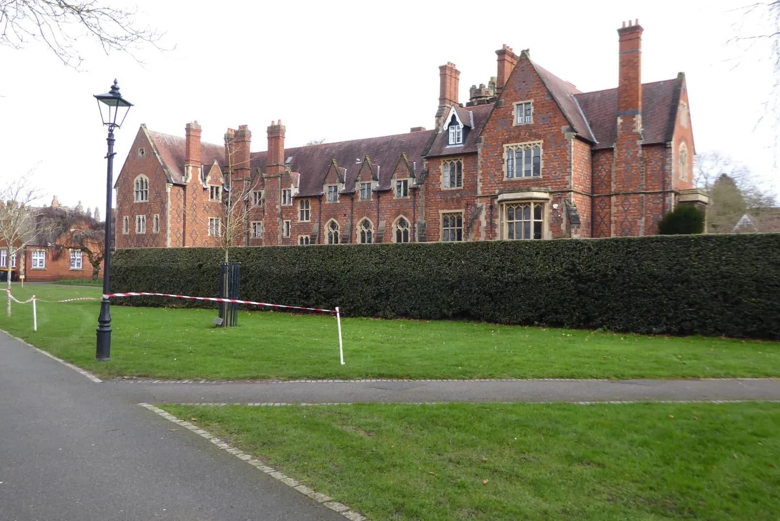 A long, red brick building with multiple gables, windows, and chimneys, sits behind a tall, neat green hedge, with a lamppost and a grassy area in the foreground.