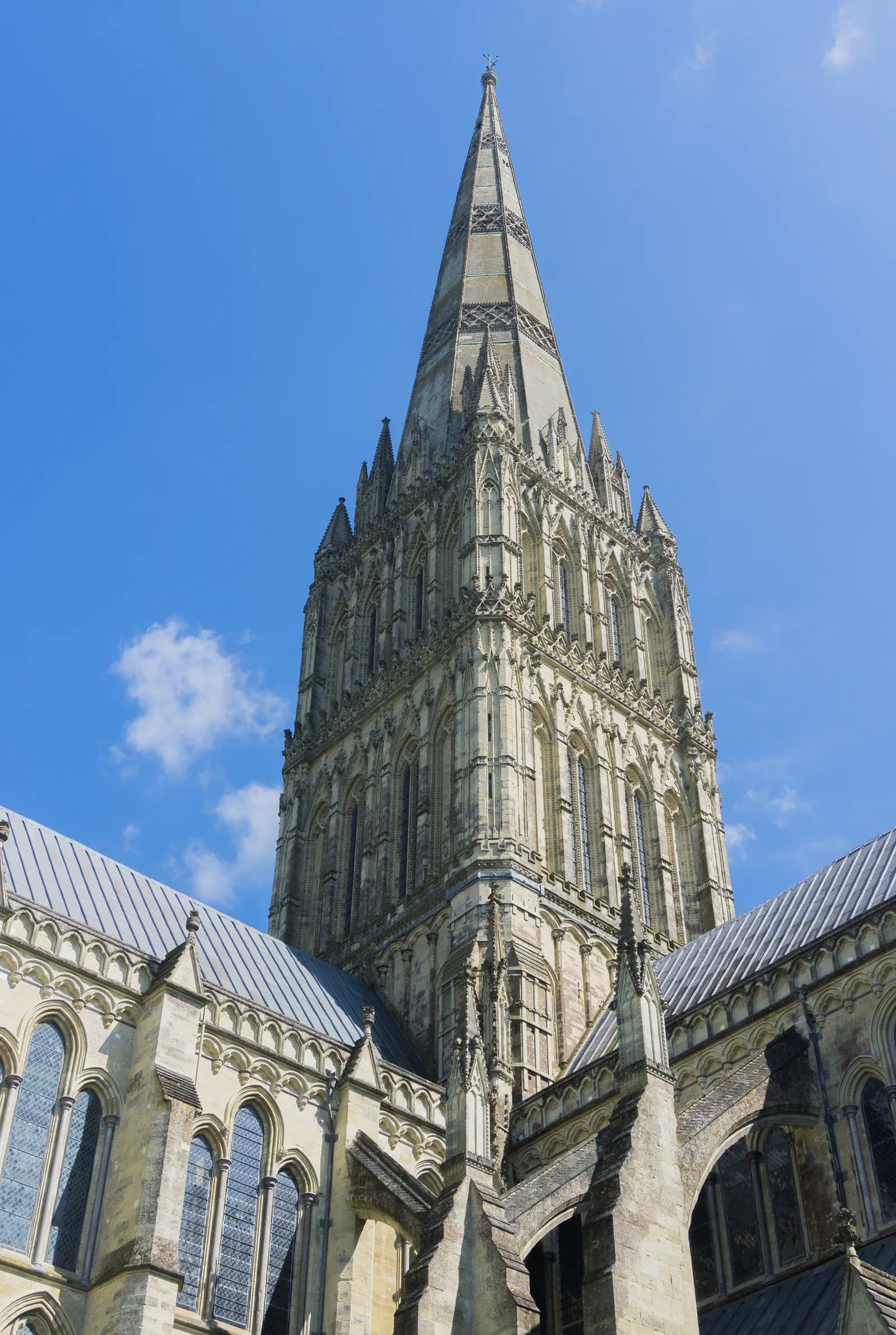 A low-angle view showcases the intricate spire and upper levels of a historic stone cathedral, contrasted against a bright blue sky with sparse clouds.