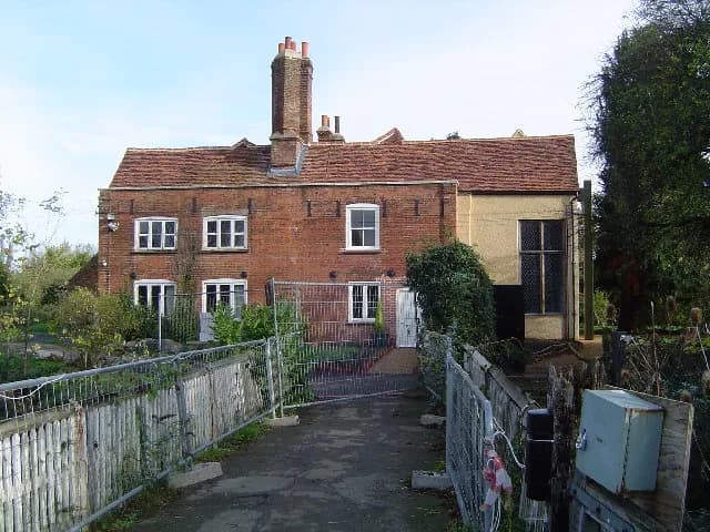A red brick house with a brown tile roof and prominent chimney stands behind a rudimentary metal and wooden fence, with some greenery around the house.
