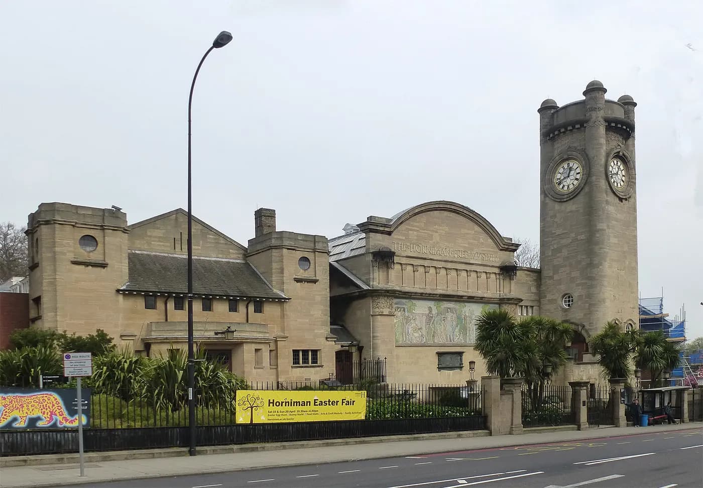 A long shot of the Horniman Museum and Gardens building complex, with a yellow banner advertising the Horniman Easter Fair on the fence in front of the museum and a clock tower on the right.