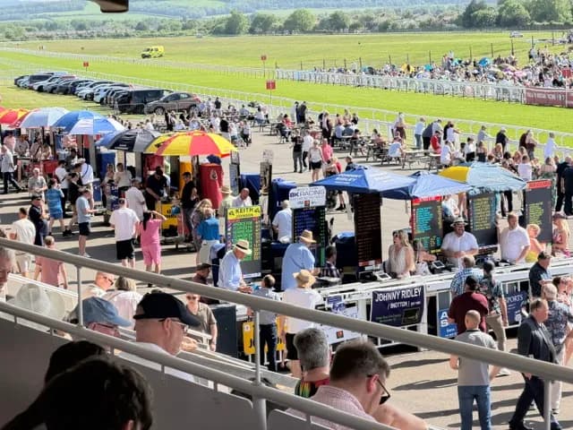 A bustling racecourse with many spectators, market stalls with colorful umbrellas, and cars parked along the track, under a clear sky.