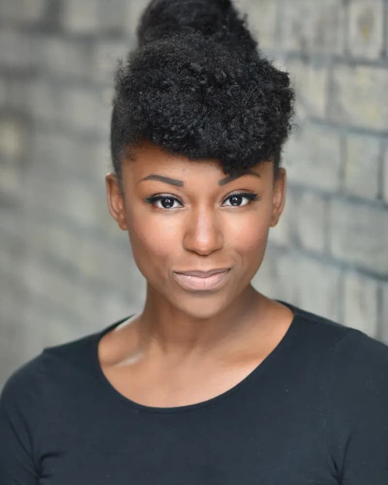 A woman with dark skin and curly black hair styled up is looking directly at the camera with a slight smile.