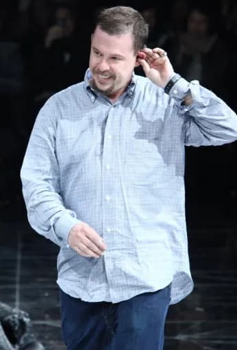 A man in a light blue button-down shirt and jeans stands, smiling while touching his right ear.