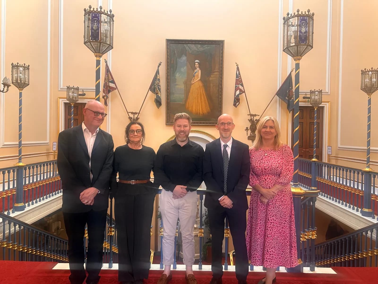 Five people, three men and two women, dressed in business or business casual attire, stand on a grand staircase in front of a painting of a woman in a yellow dress.