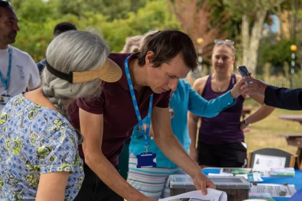 A man with a blue lanyard looks down at papers on a table, while other people, including a woman in a visor and another woman holding a phone, are also present in an outdoor setting.