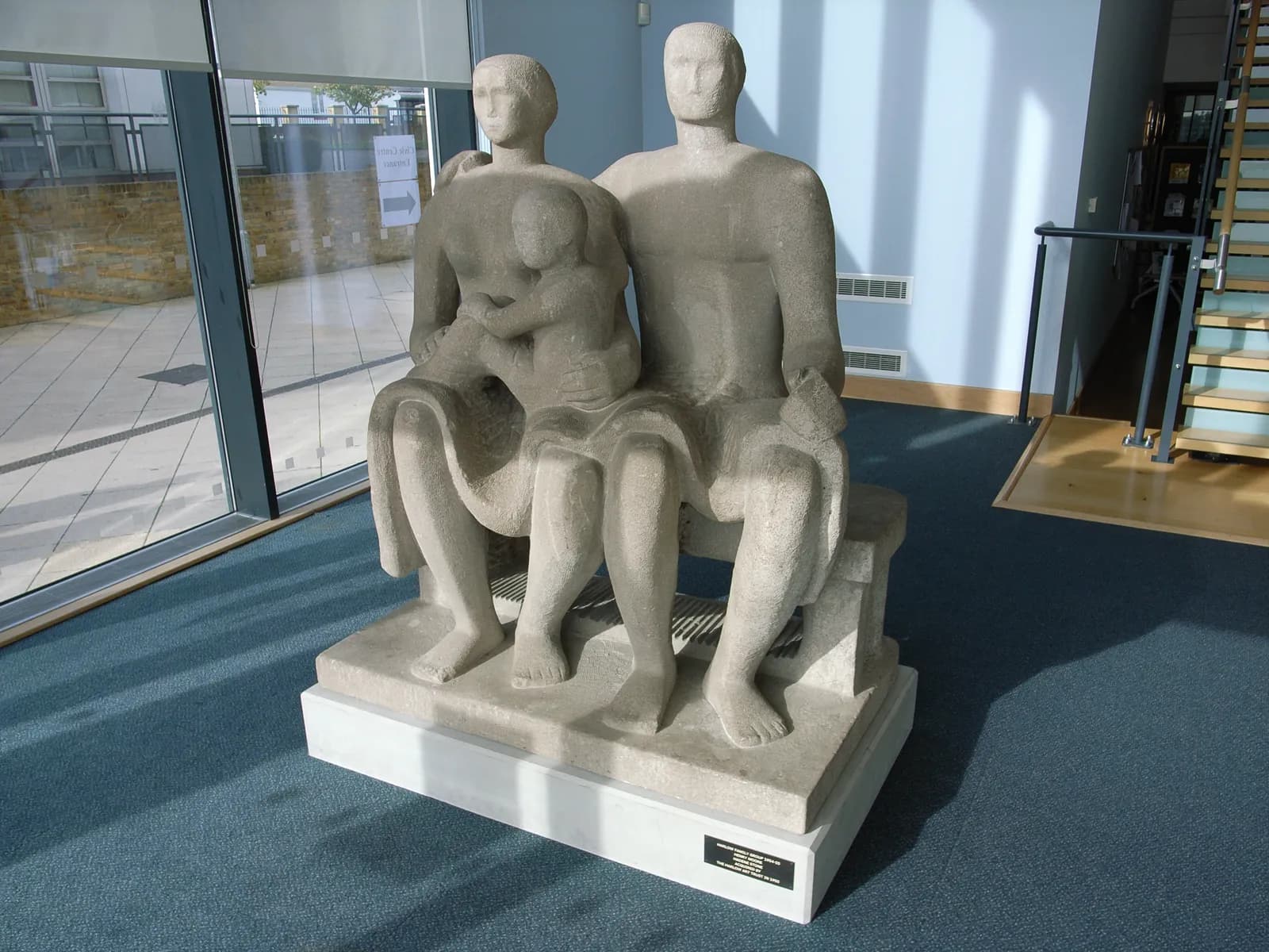 A light-colored stone sculpture shows two seated adults, a man and a woman, with a small child sitting in the woman's lap, all together on a bench.