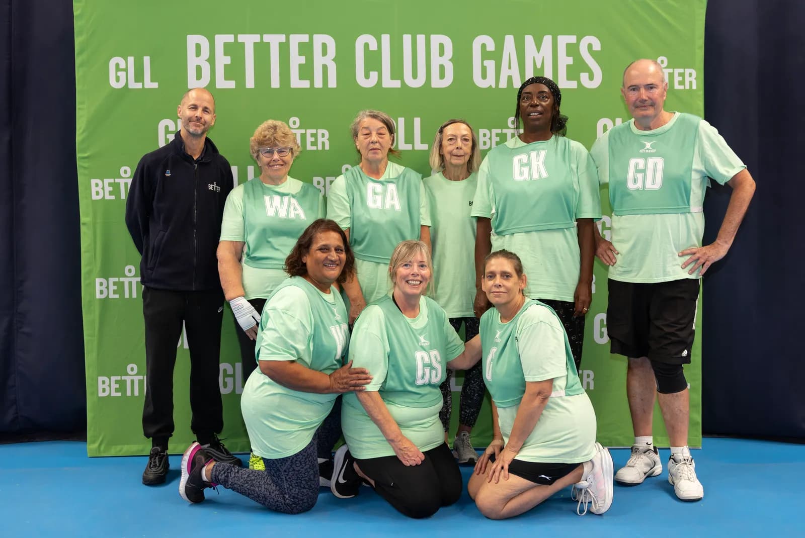 A group of older adults and one younger man in sports attire pose for a team photo in front of a green banner that says GLL Better Club Games.