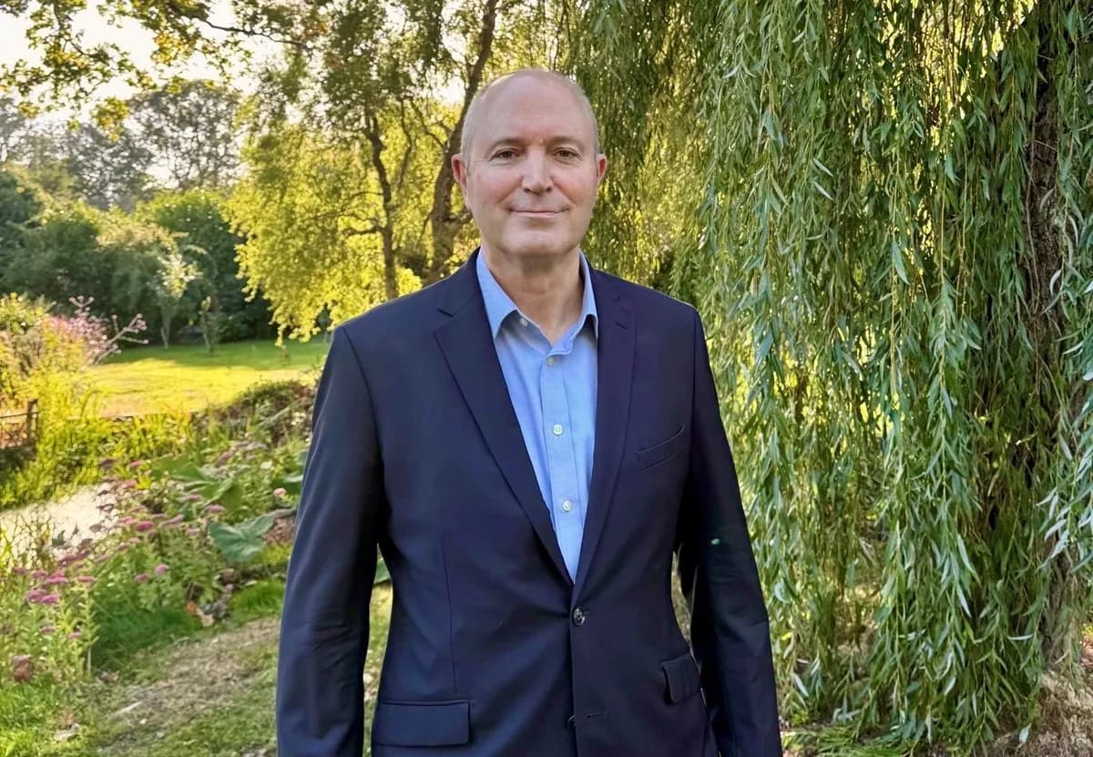 A man in a blue shirt and dark suit jacket stands outdoors, smiling softly at the viewer with green foliage and a weeping willow in the background.