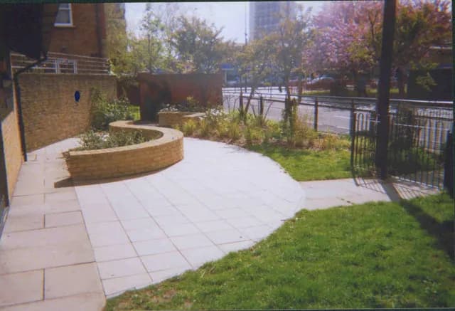 A newly landscaped area features a curved brick planter filled with shrubs, light-colored paving stones, and a grassy lawn next to a street with a black metal fence.