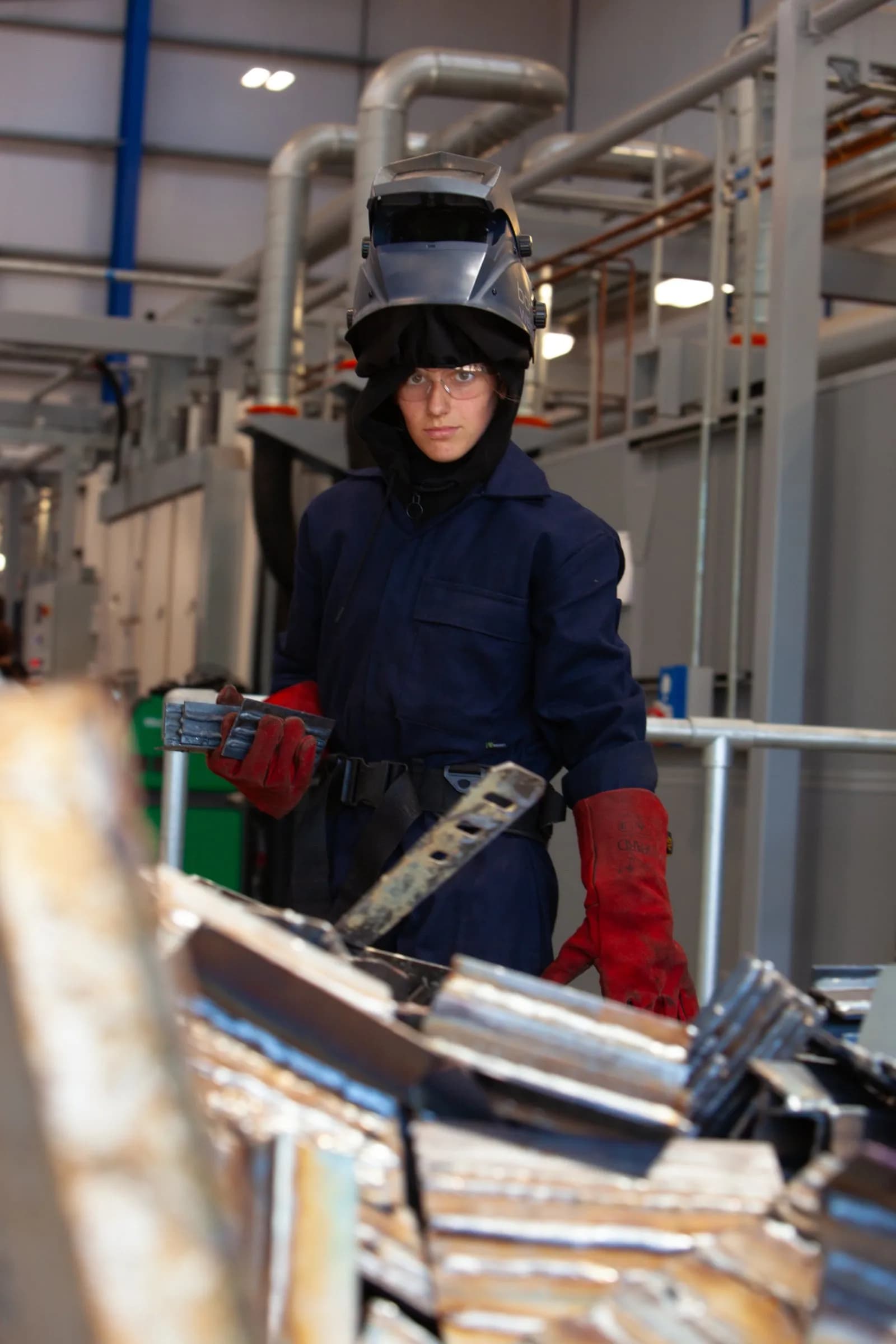 A welder wearing a dark blue jumpsuit, red gloves, safety goggles, and a welding helmet stands in front of industrial machinery, holding metal pieces in their left hand.