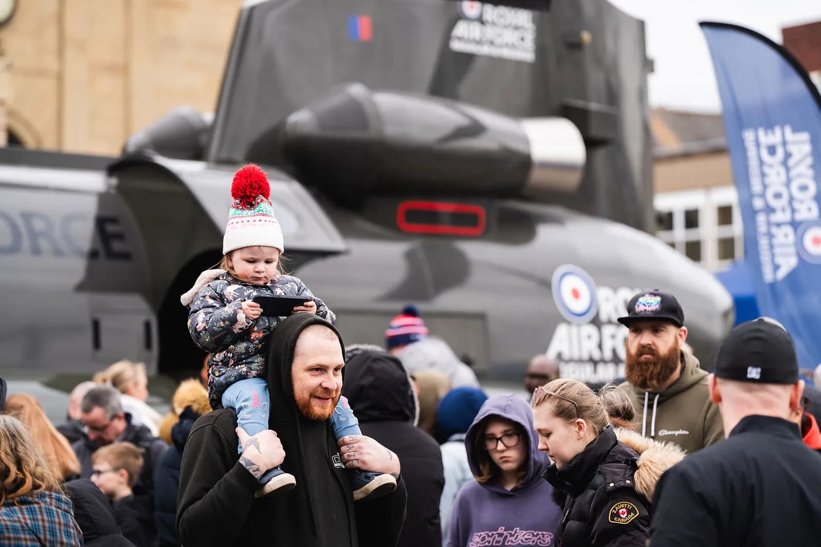 A man carries a young child on his shoulders, both looking at a phone, with a Royal Air Force helicopter and other spectators in the background.