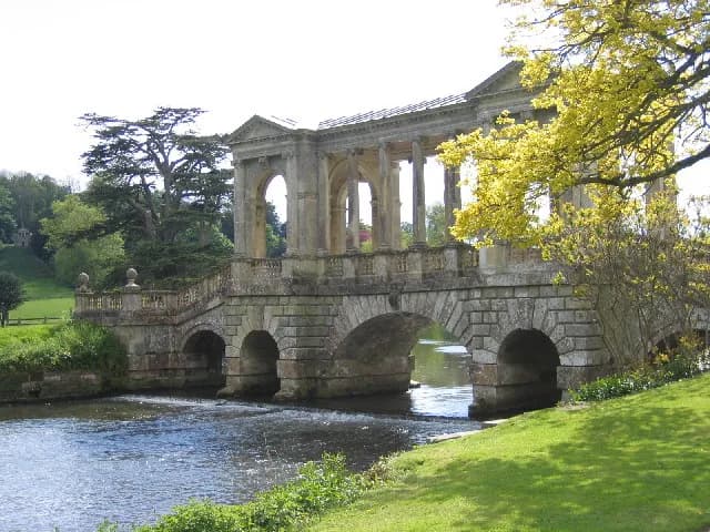 A large, ornate stone bridge with archways spans a flowing river, with green grass and trees on either bank under a bright sky.
