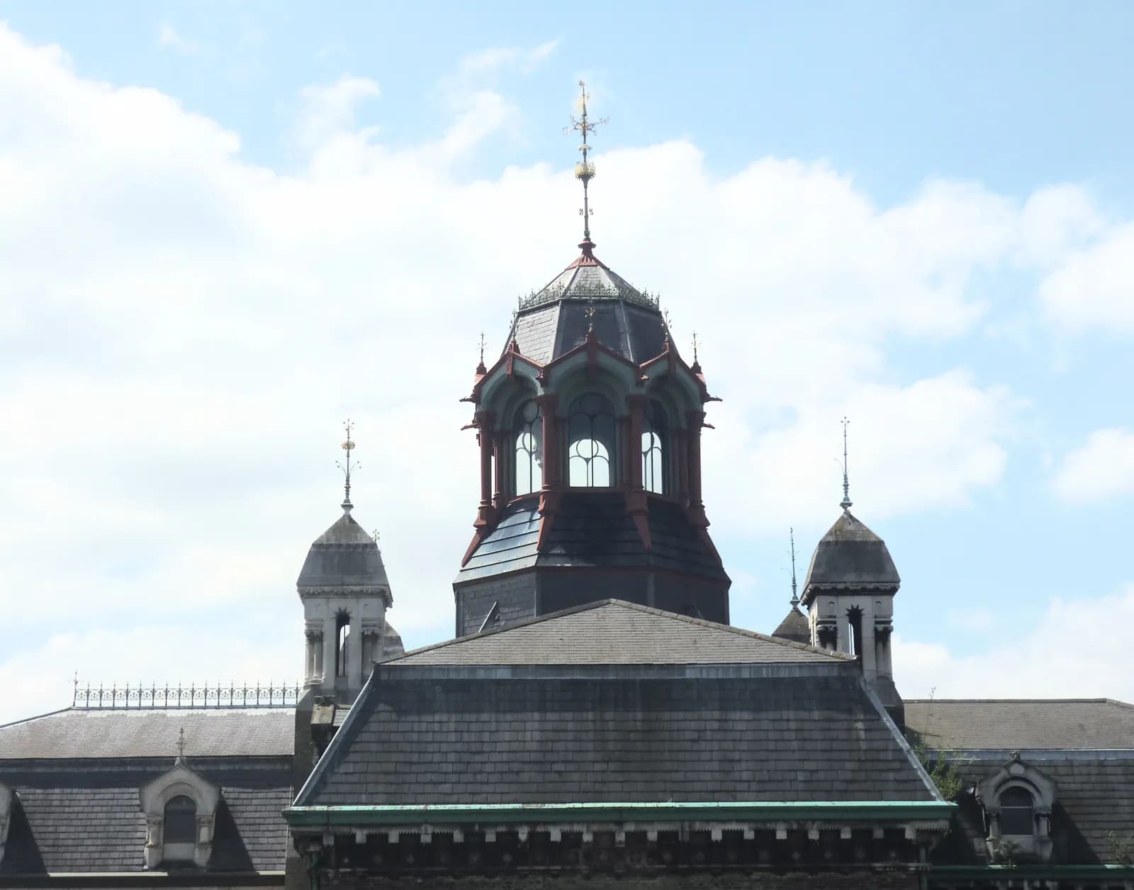 A close-up view of the intricate details on the roof of a large, old building, featuring multiple dome-like structures and a prominent octagonal tower with arched windows, set against a bright, cloudy sky.