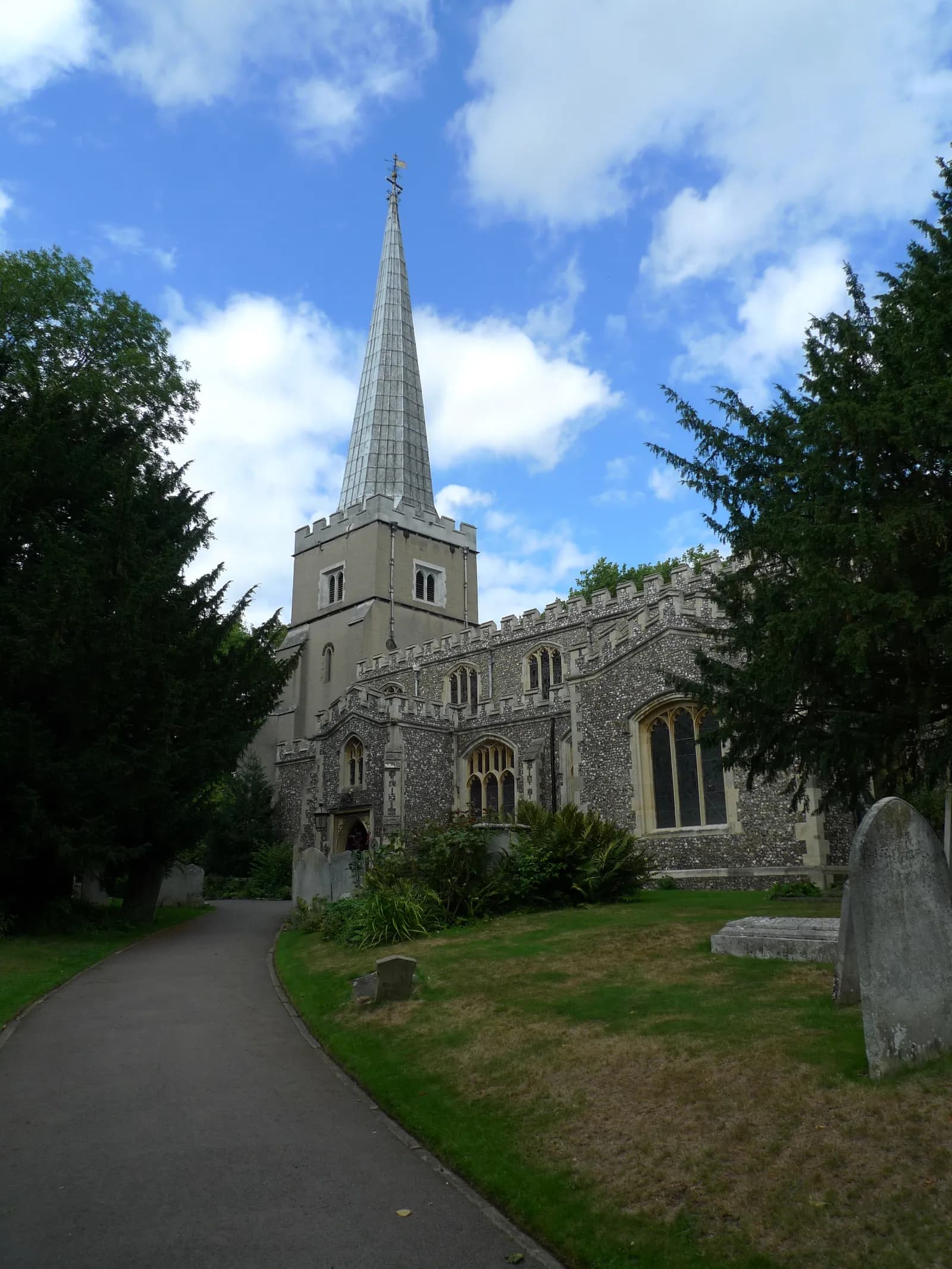 A grand, grey stone church with a tall, pointed steeple stands under a blue sky with fluffy white clouds, surrounded by green grass and trees, with a winding pathway leading towards it.