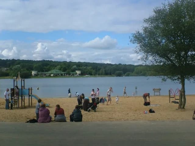 A sandy beach with a playground and people enjoying the day, with a lake and tree-lined shore in the background under a partly cloudy sky.