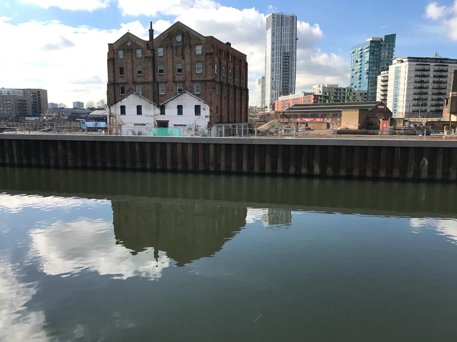 A historic brick warehouse and two smaller white buildings are reflected in the calm water of a river or canal, with modern high-rise buildings in the background under a partly cloudy sky.