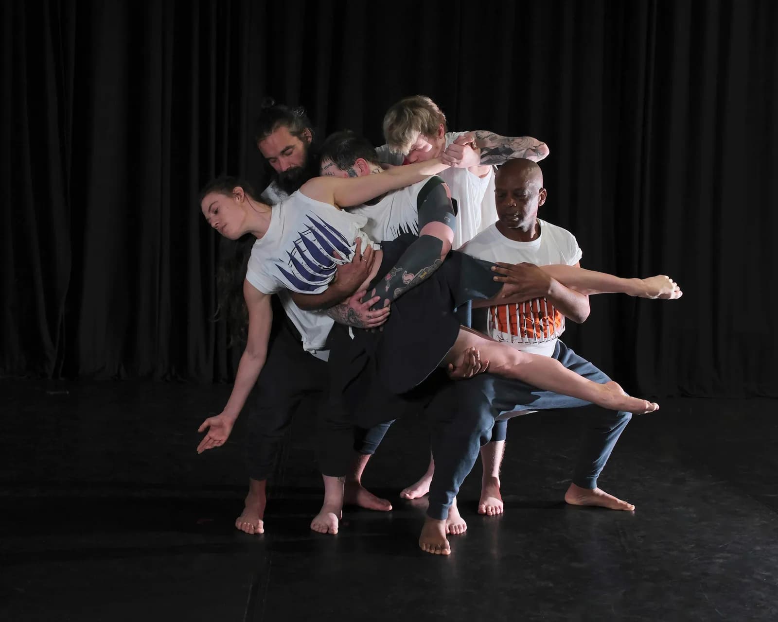 A group of five dancers, some men and one woman, are intertwined in a complex dance pose, all wearing casual clothes with bare feet against a dark background.