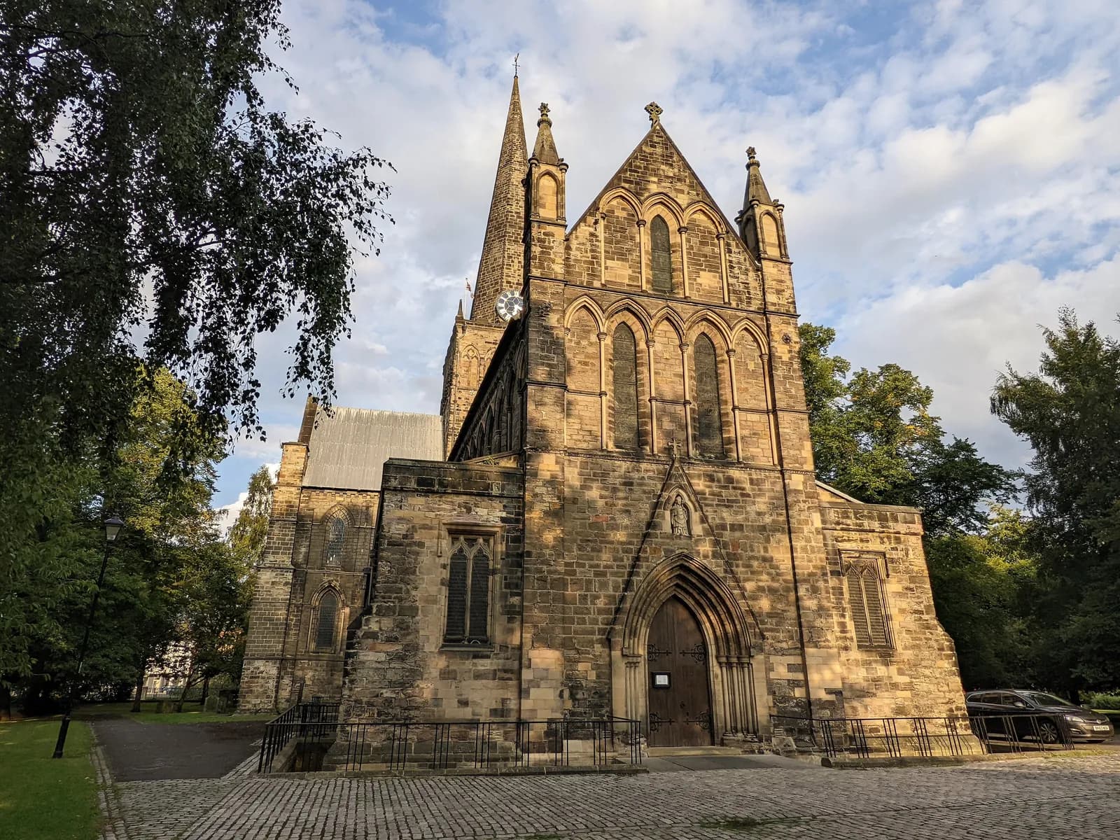 A historic stone church with multiple spires and arched windows stands under a partly cloudy sky, surrounded by green trees and a paved walkway.