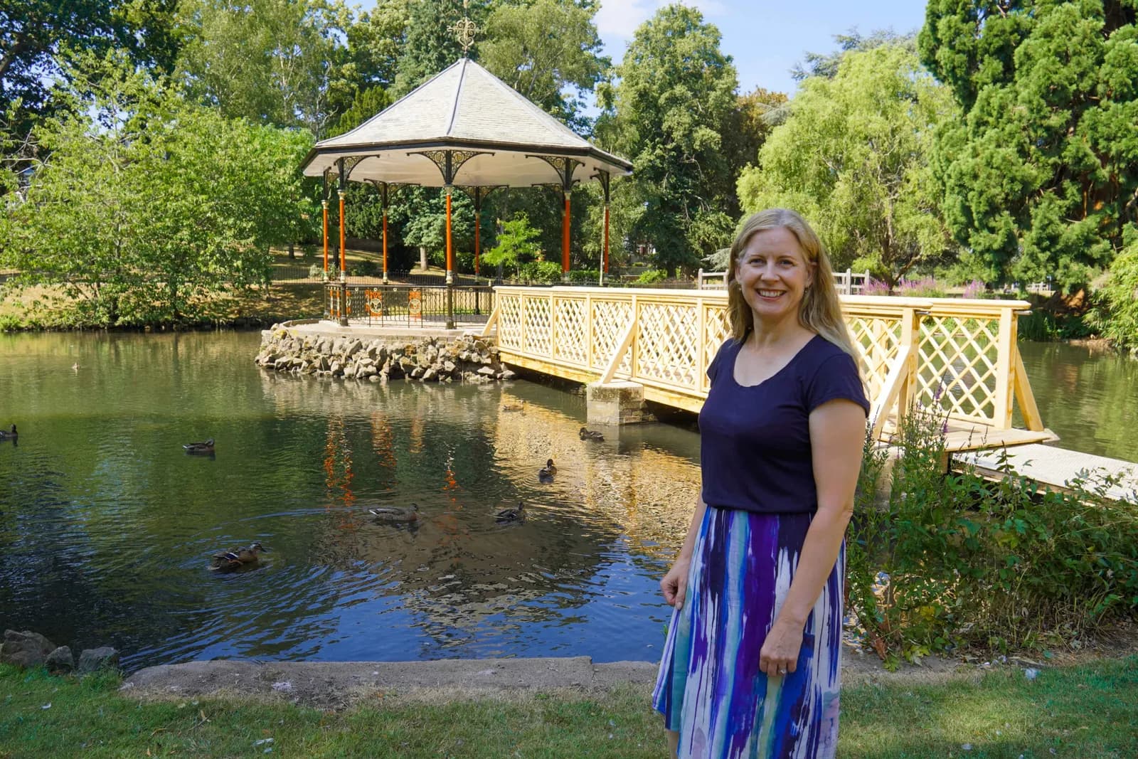 A smiling woman with blonde hair stands to the right of a pond featuring a gazebo and a wooden bridge, surrounded by lush green trees.