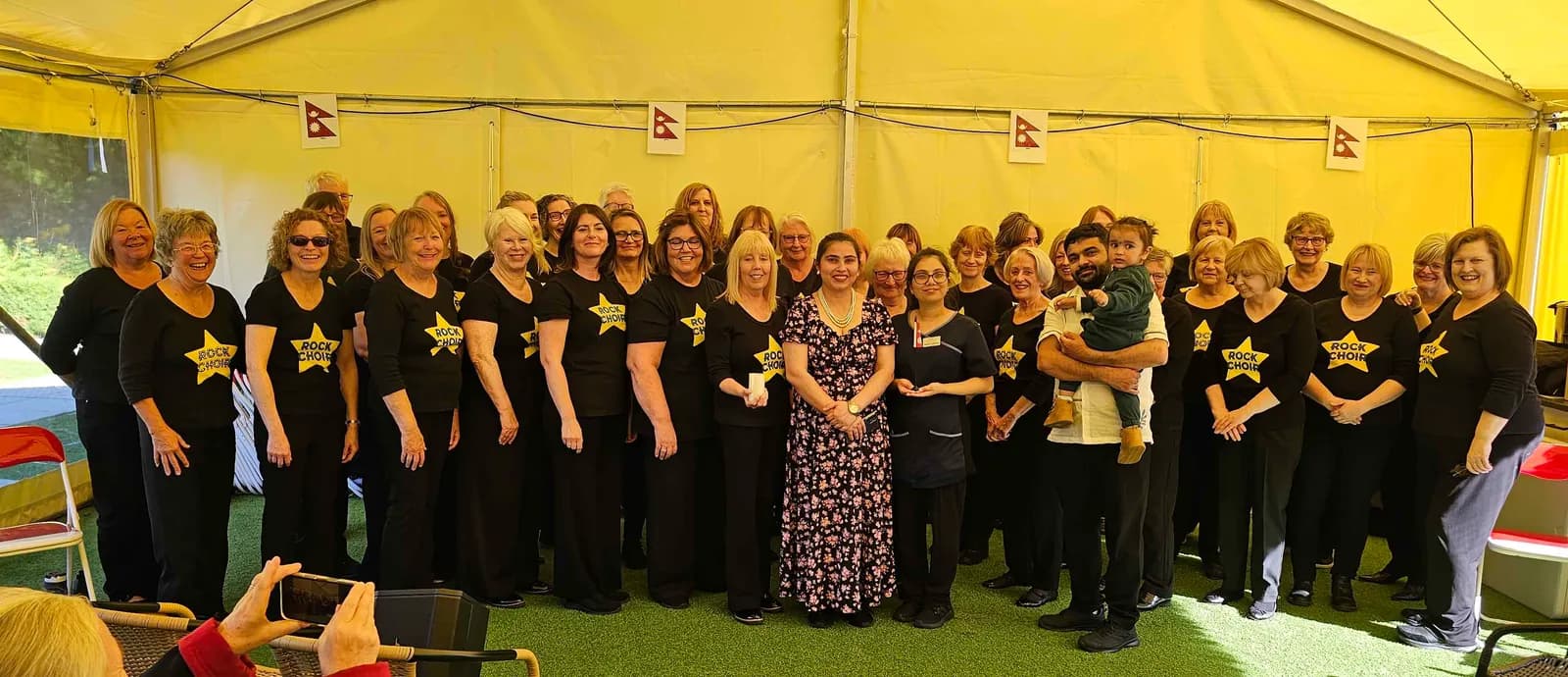 A large group of smiling adults, mostly women, wearing black shirts with a yellow star and "ROCK CHOIR" text, gathered for a group photo under a yellow tent, with one man holding a small child and another woman wearing a floral dress.