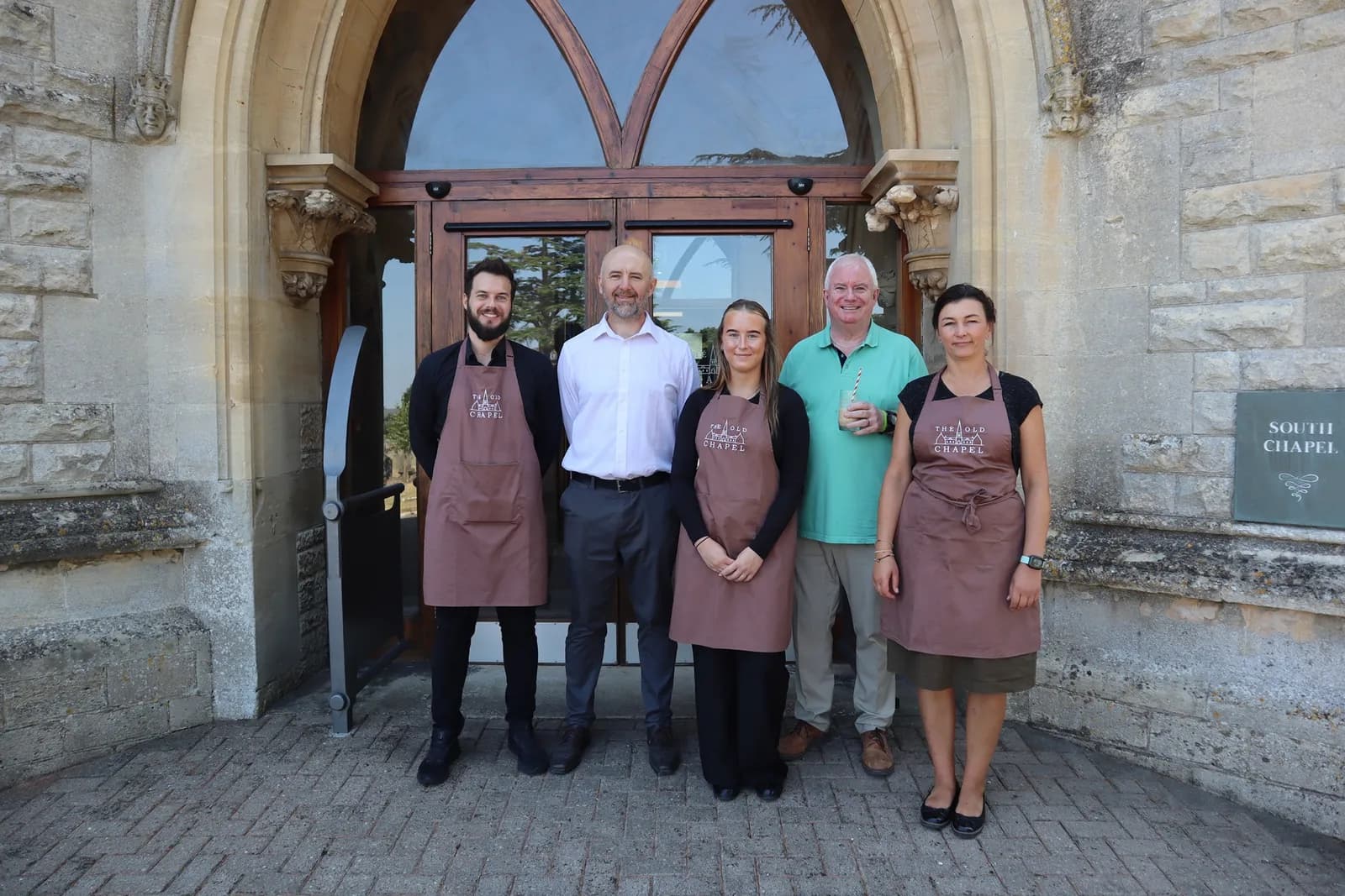 A group of five people, two men and a woman in brown aprons, a man in a white shirt, and a man in a green polo shirt, stand smiling in front of a large wooden door with arched windows of a stone building.