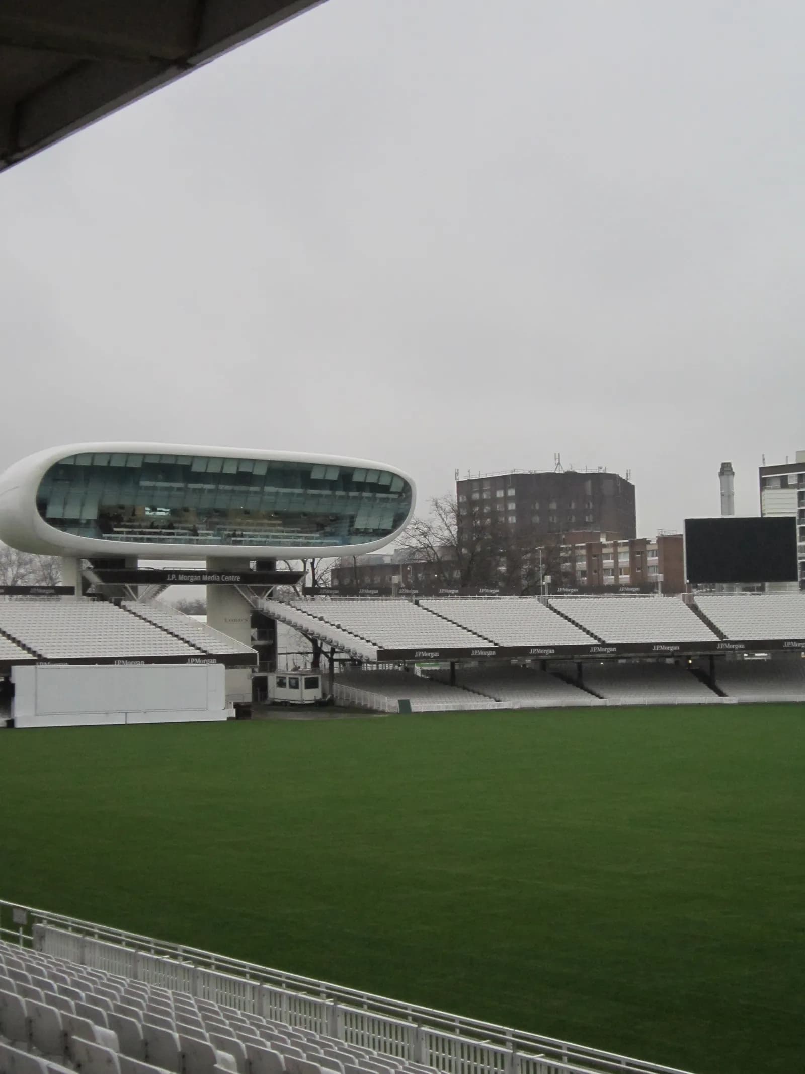 A large, modern, white building with extensive windows overlooks a bright green grass field, surrounded by rows of empty white stadium seating under a cloudy sky.