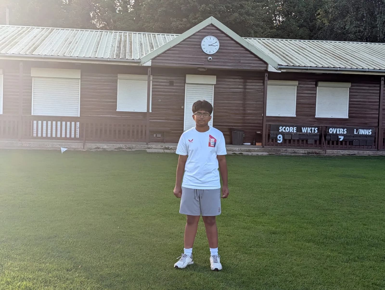A young boy wearing a white t-shirt and grey shorts stands on a green grass field in front of a wooden building with a score board.
