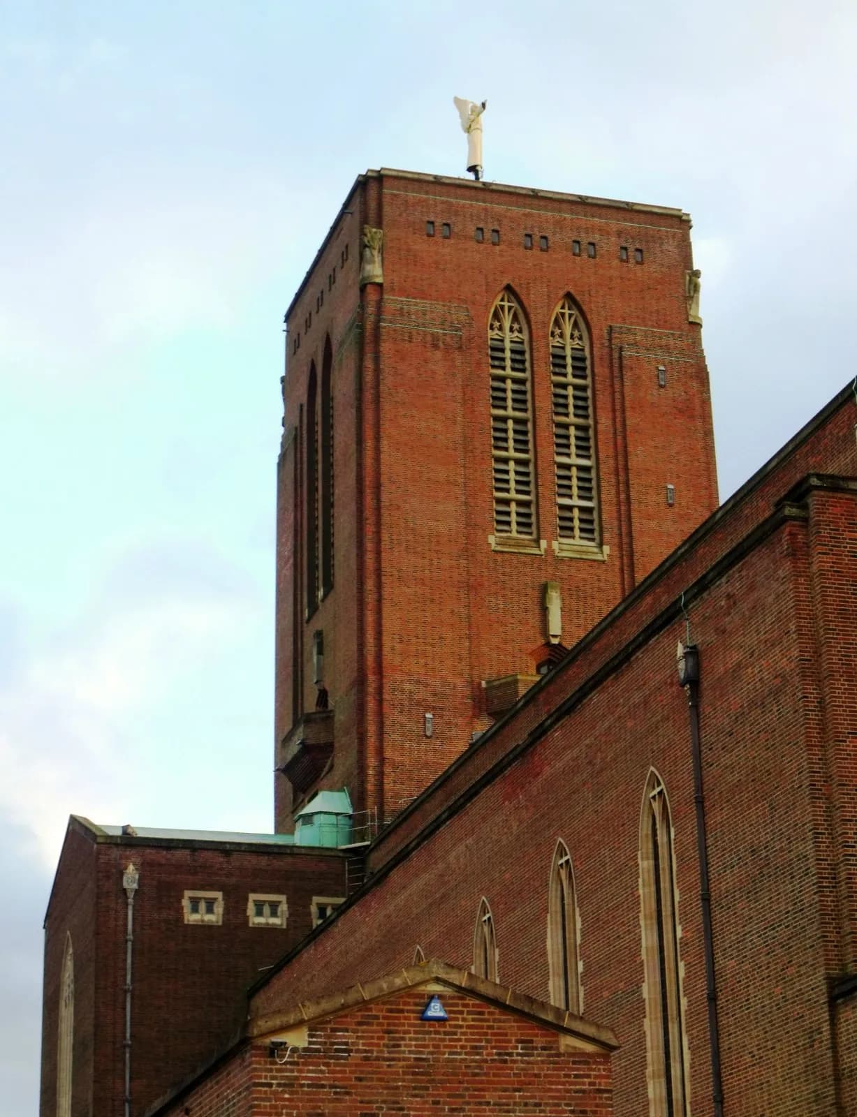 A tall brick church tower with a white statue of a robed figure on top reaches into a cloudy sky, with parts of the church's brick facade in the foreground.