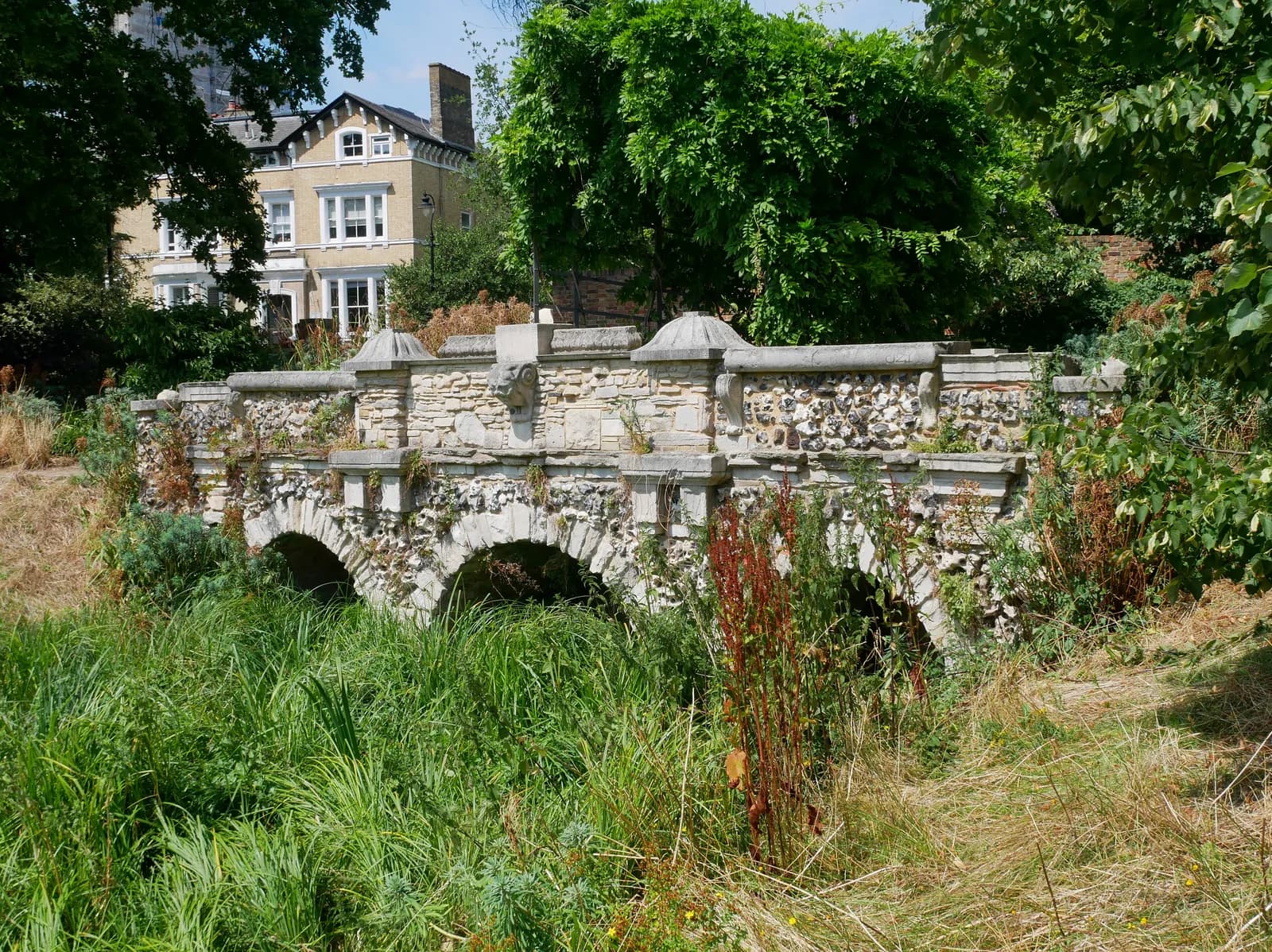 A stone bridge with multiple arches stands over thick green foliage, with a large house visible behind trees in the background.